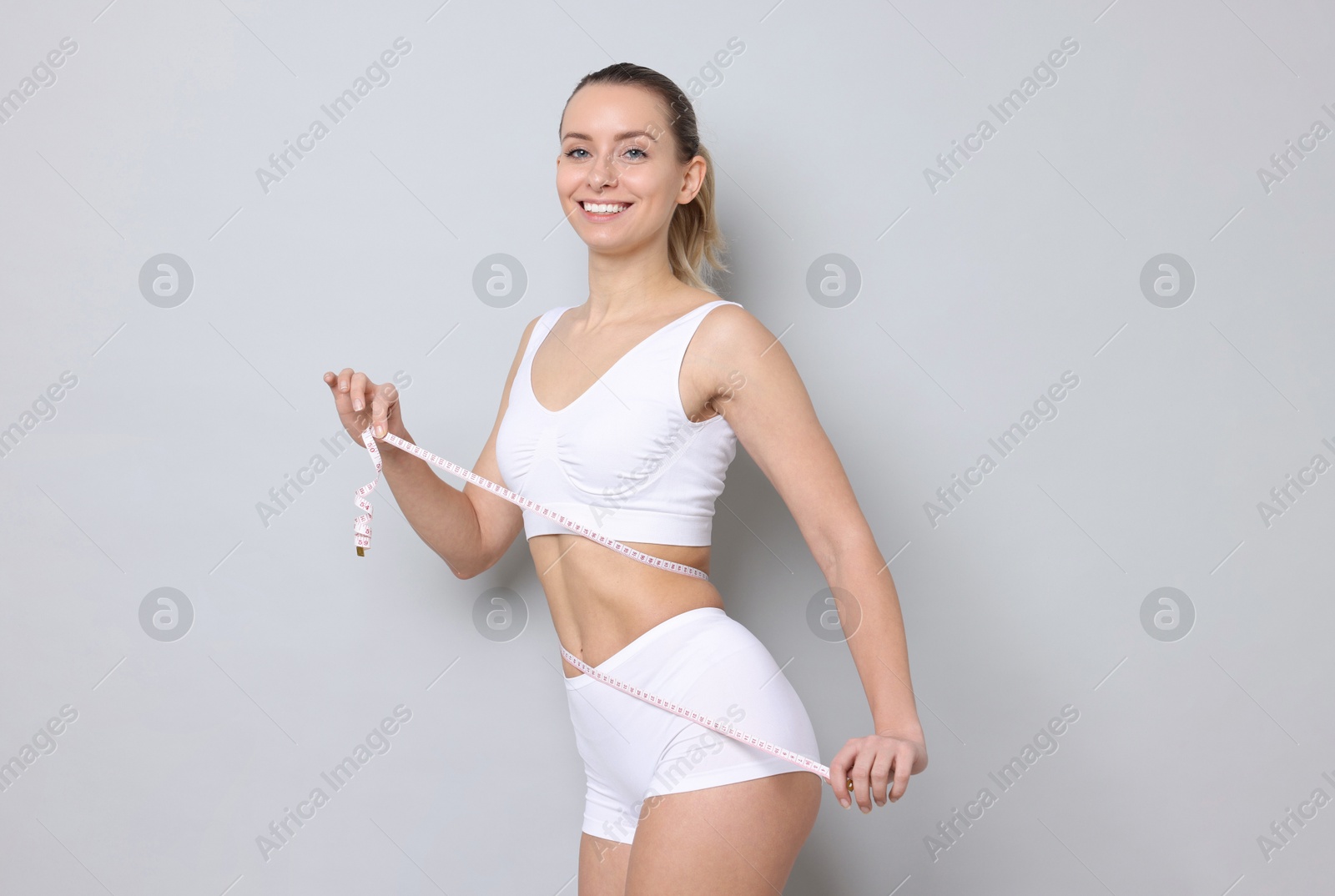 Photo of Weight loss. Happy woman measuring her slim waist with tape on light grey background