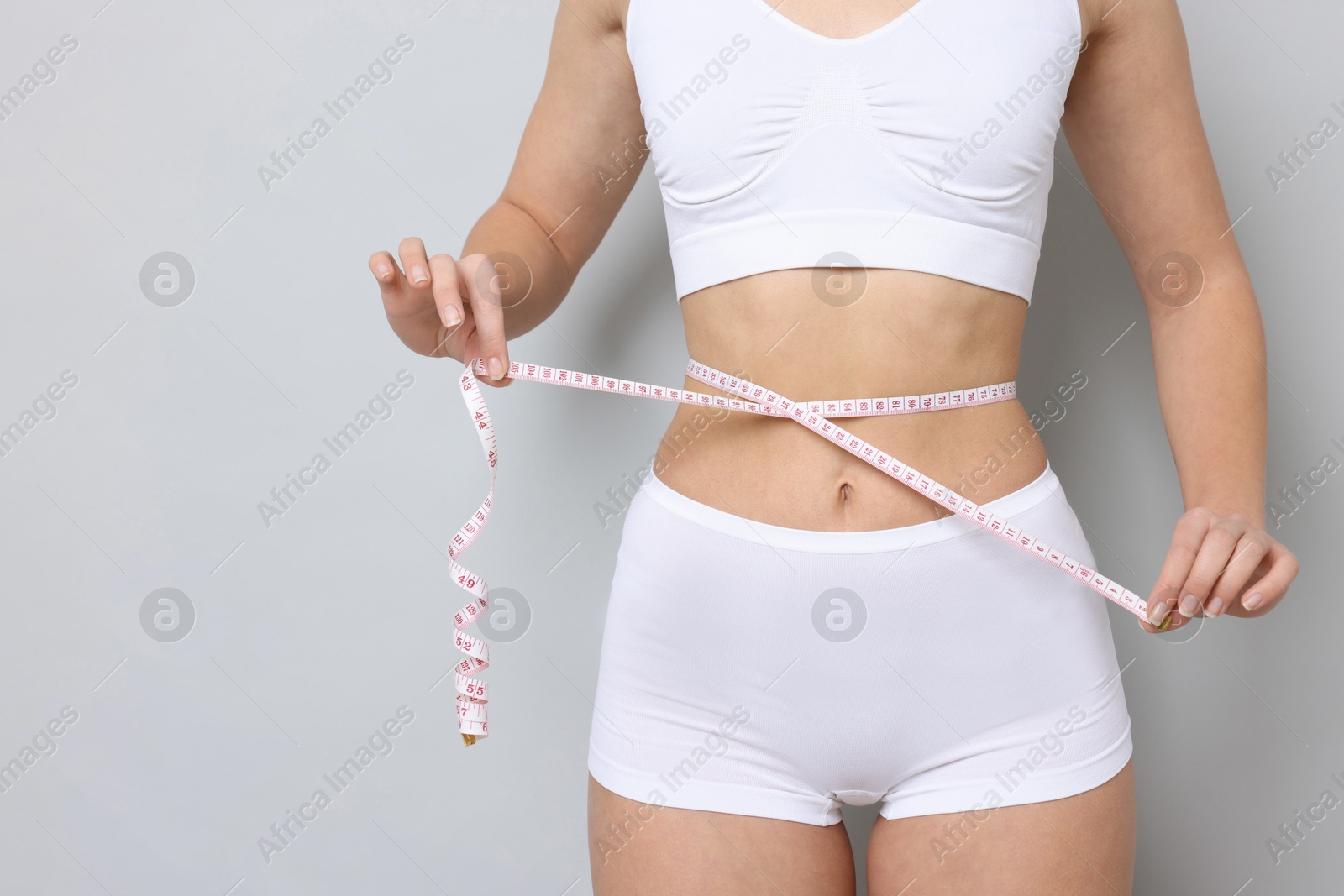 Photo of Weight loss. Woman measuring her slim waist with tape on light grey background, closeup. Space for text