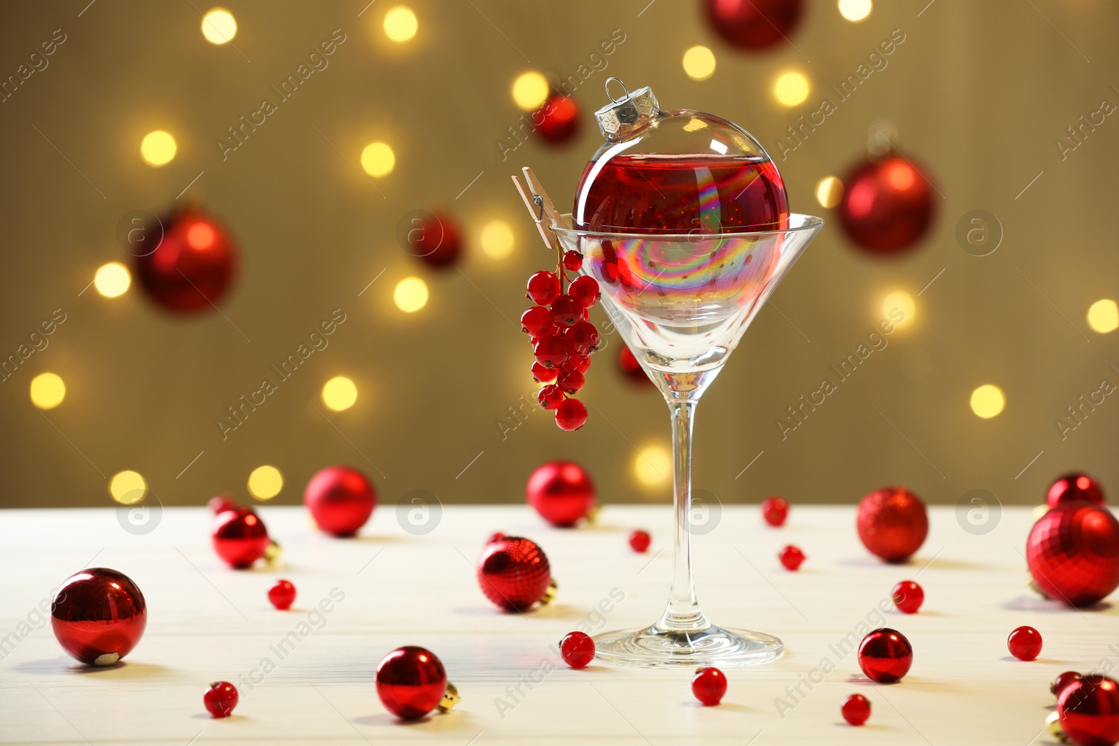 Photo of Creative presentation of Christmas cocktail in bauble and glass on white table against dark beige background with blurred lights