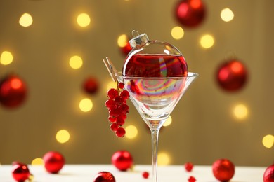 Photo of Creative presentation of Christmas cocktail in bauble and glass against dark beige background with blurred lights, closeup