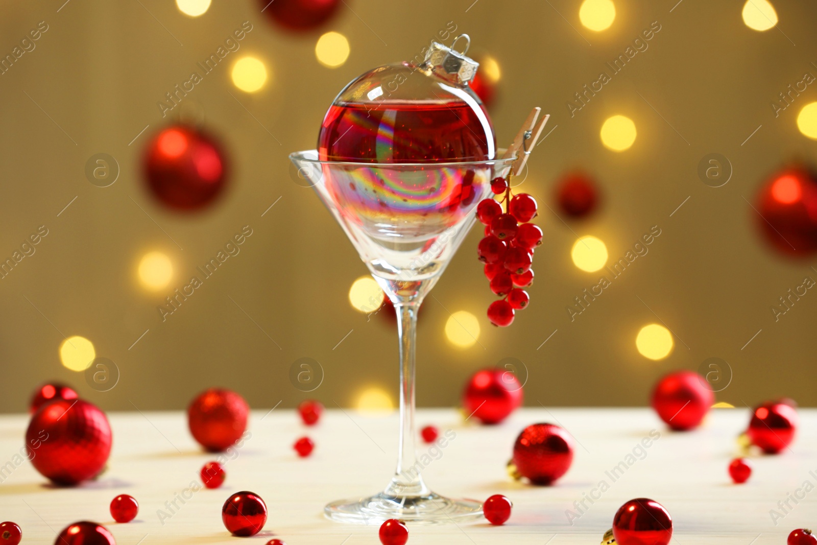 Photo of Creative presentation of Christmas cocktail in bauble and glass on white table against dark beige background with blurred lights