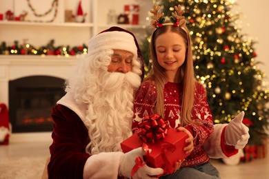 Photo of Christmas celebration. Santa Claus giving gift box to girl at home