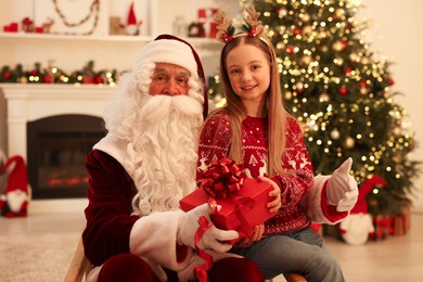 Photo of Christmas celebration. Santa Claus giving gift box to girl at home