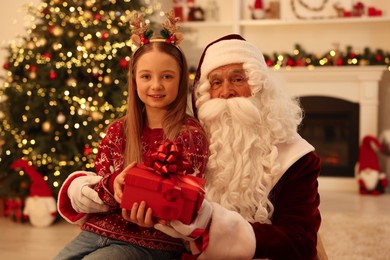 Photo of Christmas celebration. Santa Claus giving gift box to girl at home