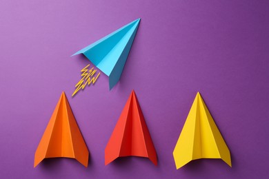 Image of Light blue paper plane bursting forward among others on purple background