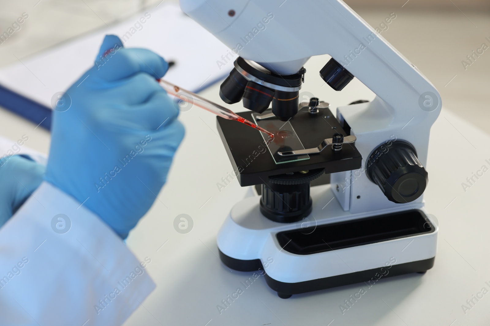 Photo of Laboratory testing. Doctor dripping blood sample onto glass slide while working with microscope at table indoors, closeup