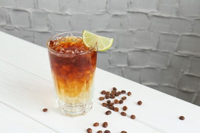 Photo of Refreshing espresso tonic drink with slice of lime and coffee beans on white wooden table. Space for text