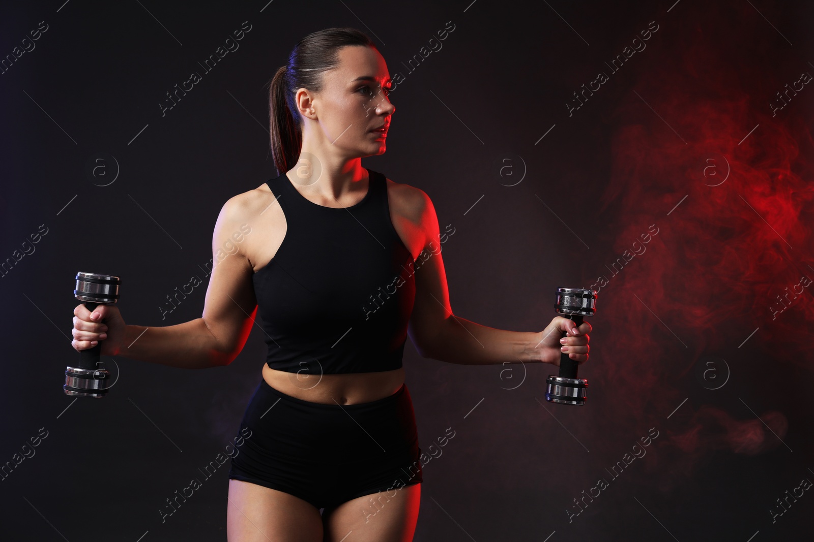 Photo of Woman exercising with dumbbells in smoke on dark background. Space for text