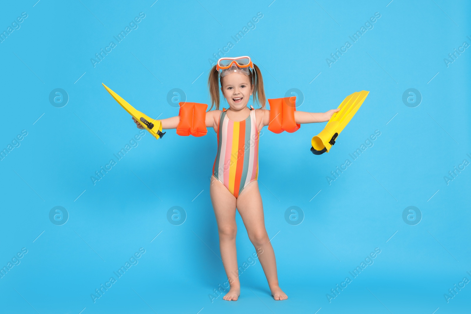 Photo of Cute little girl in swimsuit with swim armbands and fins on light blue background