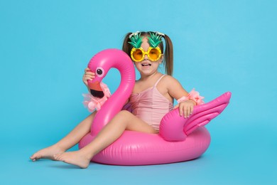 Photo of Cute little girl in swimsuit with funny sunglasses and inflatable ring on light blue background