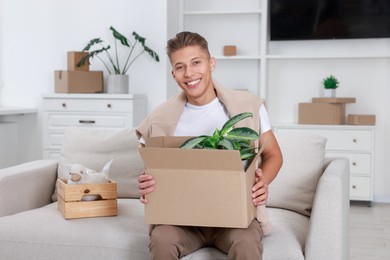 Photo of Happy man holding moving box with houseplant in new apartment. Housewarming party