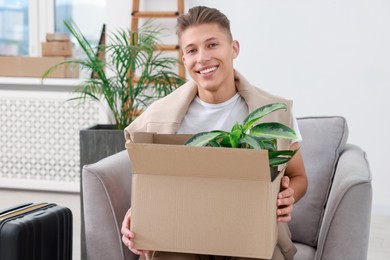 Photo of Happy man holding moving box with houseplant in new apartment. Housewarming party