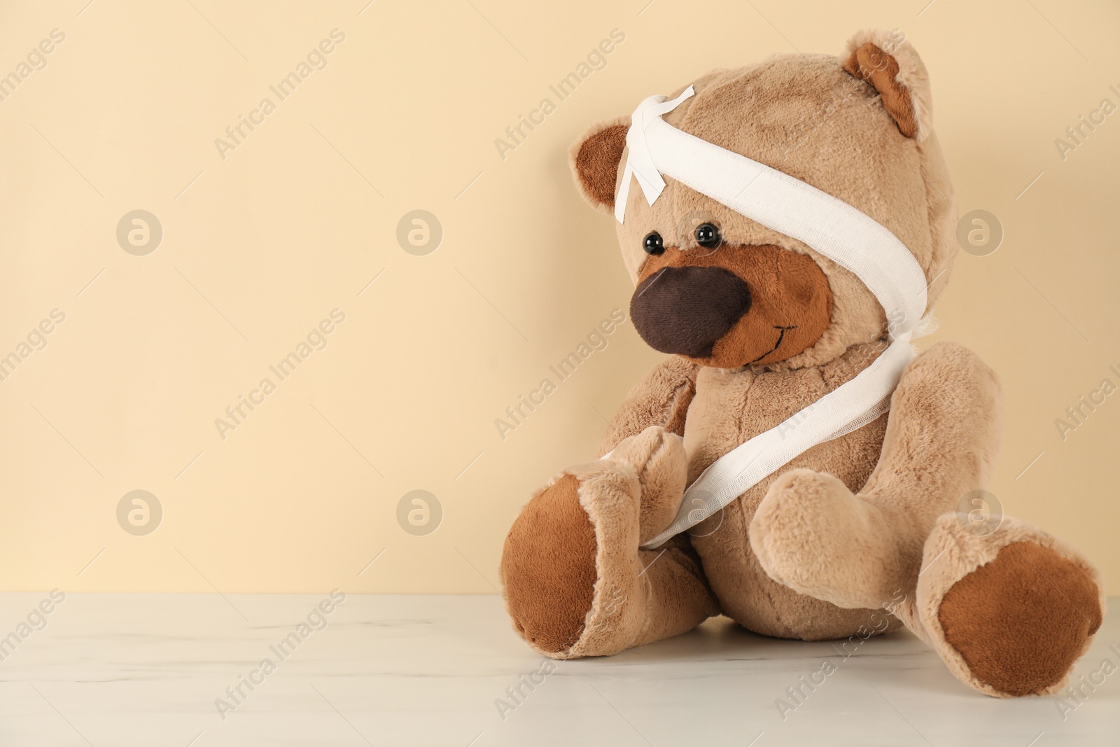 Photo of Teddy bear with bandages and sticking plasters on white marble table against beige background, space for text