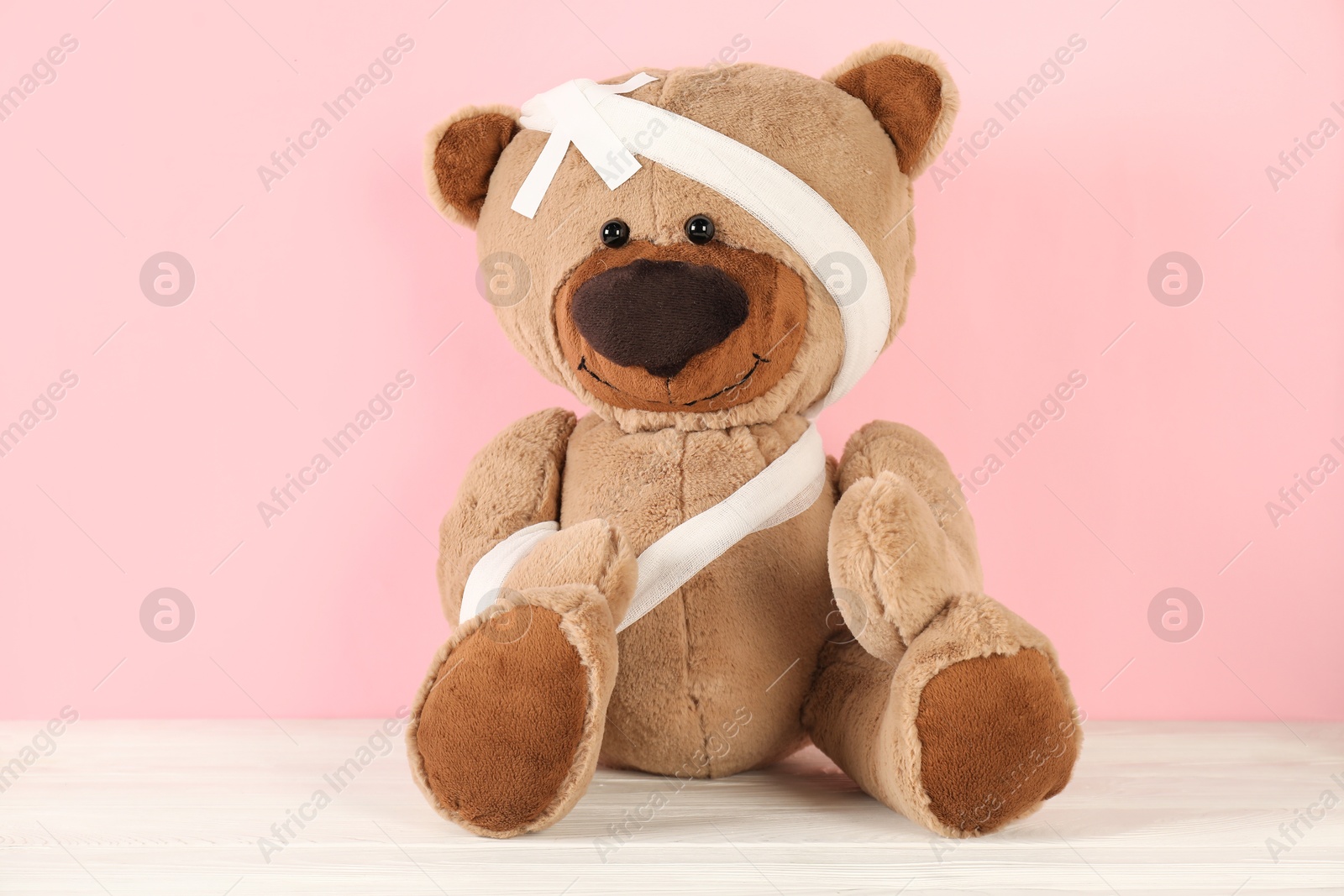 Photo of Teddy bear with bandages and sticking plasters on white wooden table against pink background