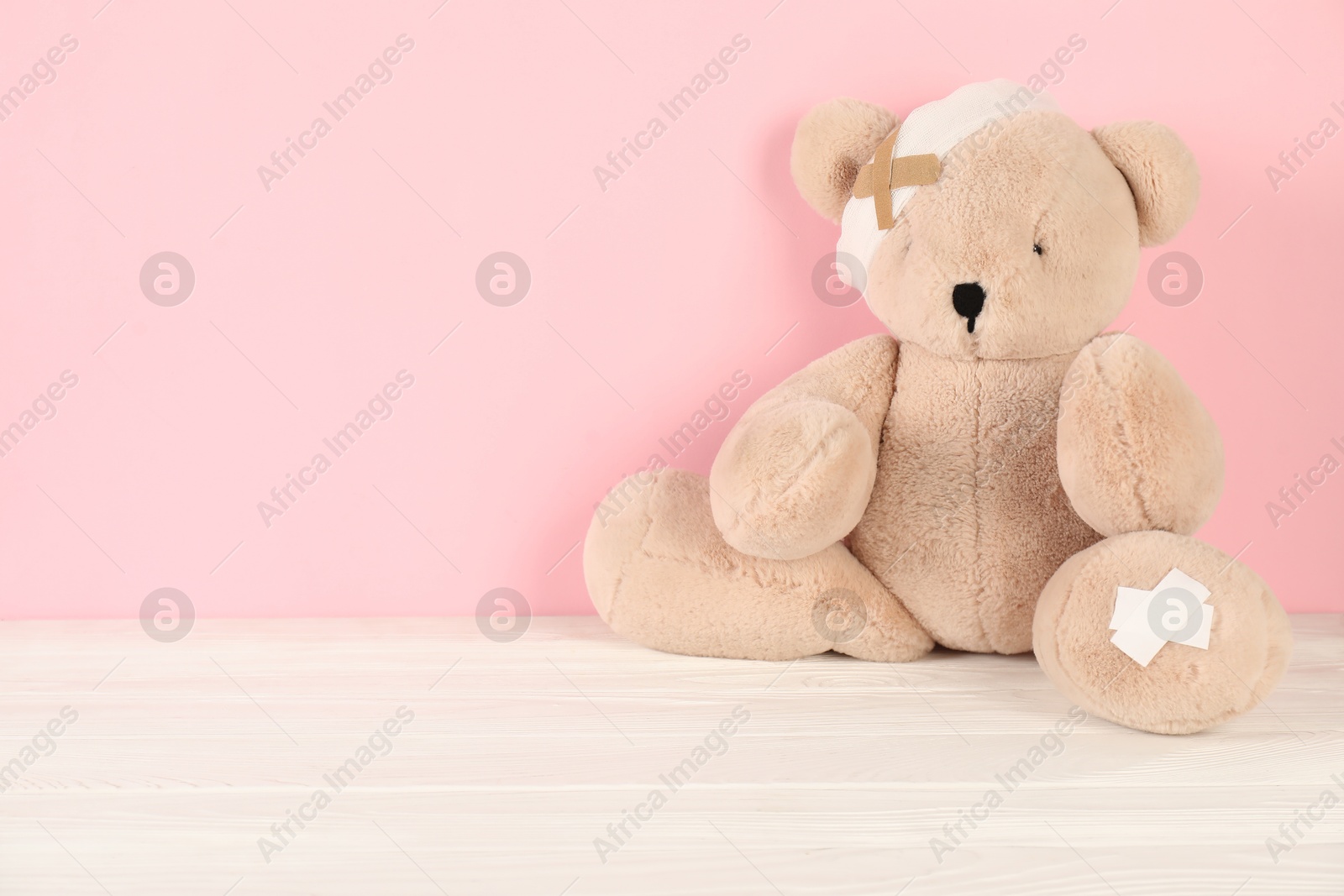 Photo of Teddy bear with bandage and sticking plasters on white wooden table against pink background, space for text