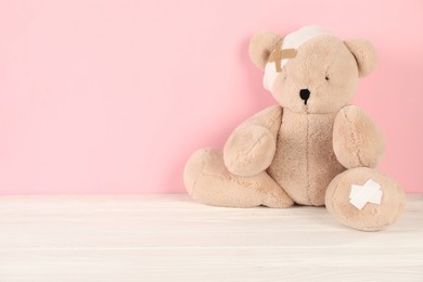 Photo of Teddy bear with bandage and sticking plasters on white wooden table against pink background, space for text