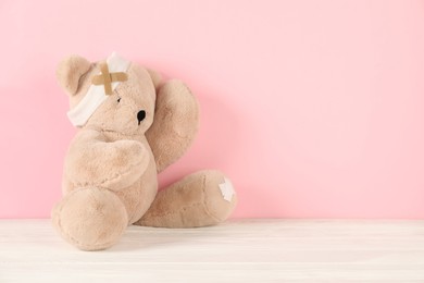 Photo of Teddy bear with bandage and sticking plasters on white wooden table against pink background, space for text