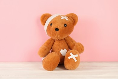 Photo of Teddy bear with bandages and sticking plasters on white wooden table against pink background