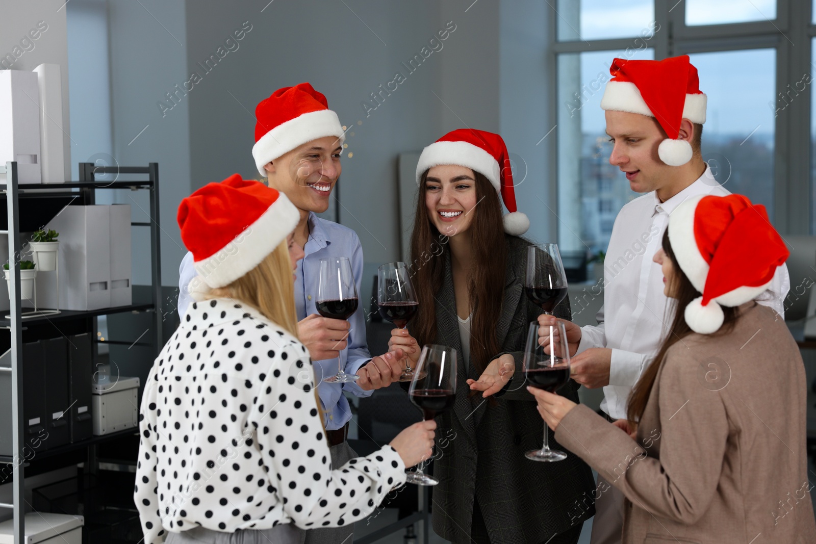 Photo of Happy coworkers in Santa hats with glasses of wine at office Christmas party