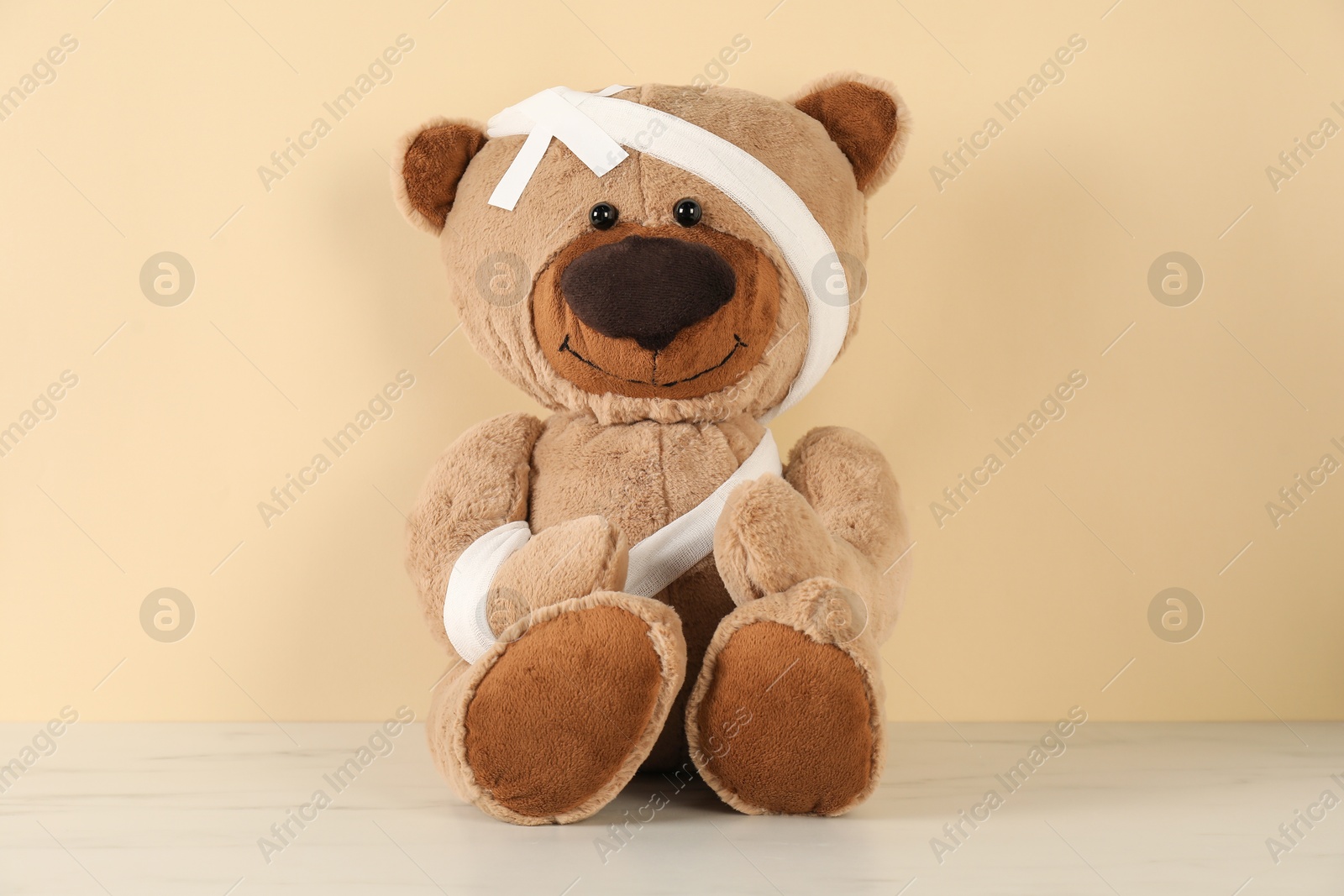 Photo of Teddy bear with bandages and sticking plasters on white marble table against beige background