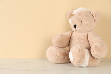 Photo of Teddy bear with bandages and sticking plasters on white marble table against beige background, space for text