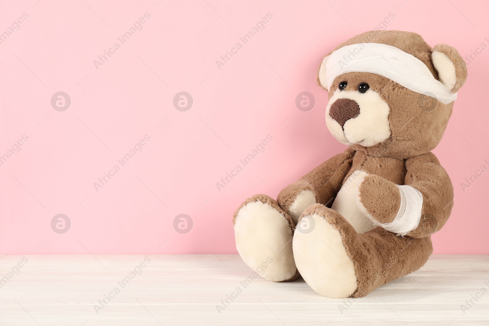 Photo of Teddy bear with bandages on white wooden table against pink background, space for text