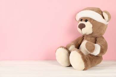 Photo of Teddy bear with bandages on white wooden table against pink background, space for text