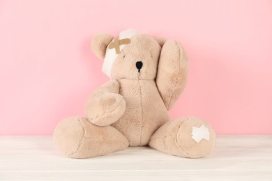 Photo of Teddy bear with bandage and sticking plasters on white wooden table against pink background