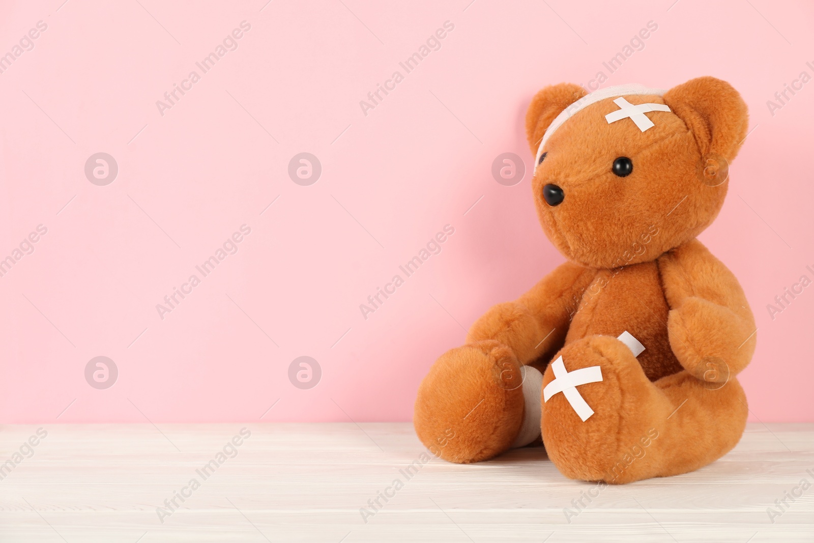 Photo of Teddy bear with bandages and sticking plasters on white wooden table against pink background, space for text