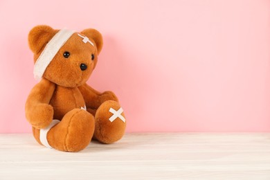 Photo of Teddy bear with bandages and sticking plasters on white wooden table against pink background, space for text