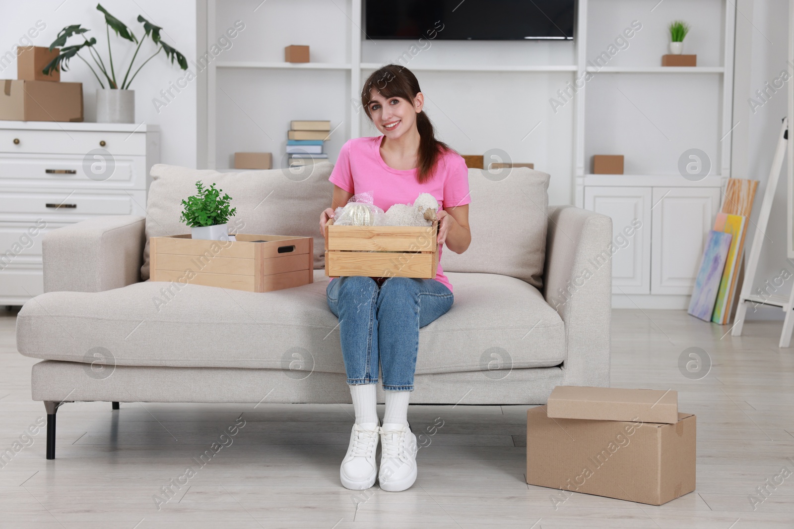 Photo of Happy woman holding wooden crate with stuff on sofa in new apartment. Housewarming party