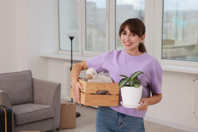 Happy woman holding wooden crate with stuff and houseplant in new apartment. Housewarming party