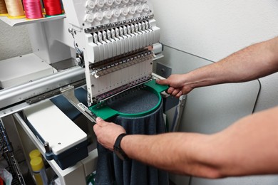 Photo of Man using embroidery machine to make design on T-shirt indoors, closeup