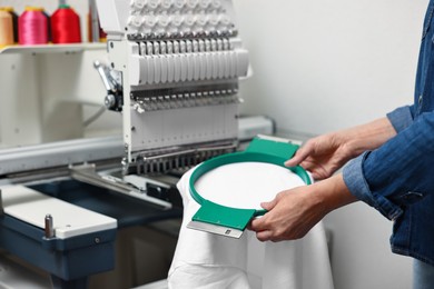 Photo of Woman using embroidery machine to make design on T-shirt indoors, closeup