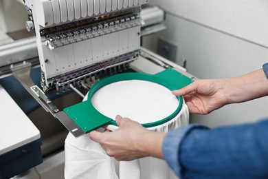 Photo of Woman using embroidery machine to make design on T-shirt indoors, closeup