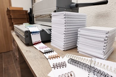 Photo of Modern binding machine, stacks of notebooks and double loop wire binding spines on wooden table indoors, closeup