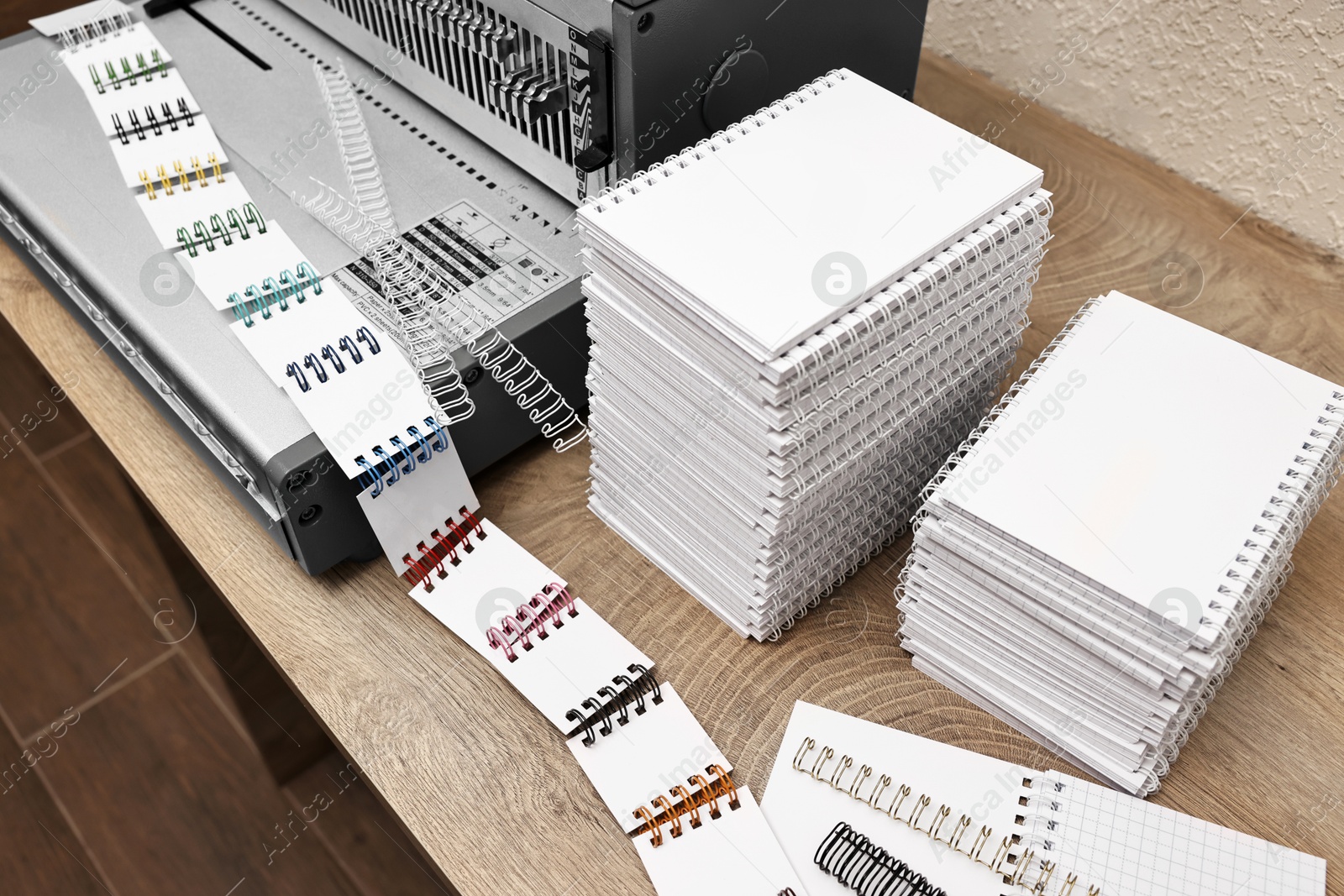 Photo of Modern binding machine, stacks of notebooks and double loop wire binding spines on wooden table indoors