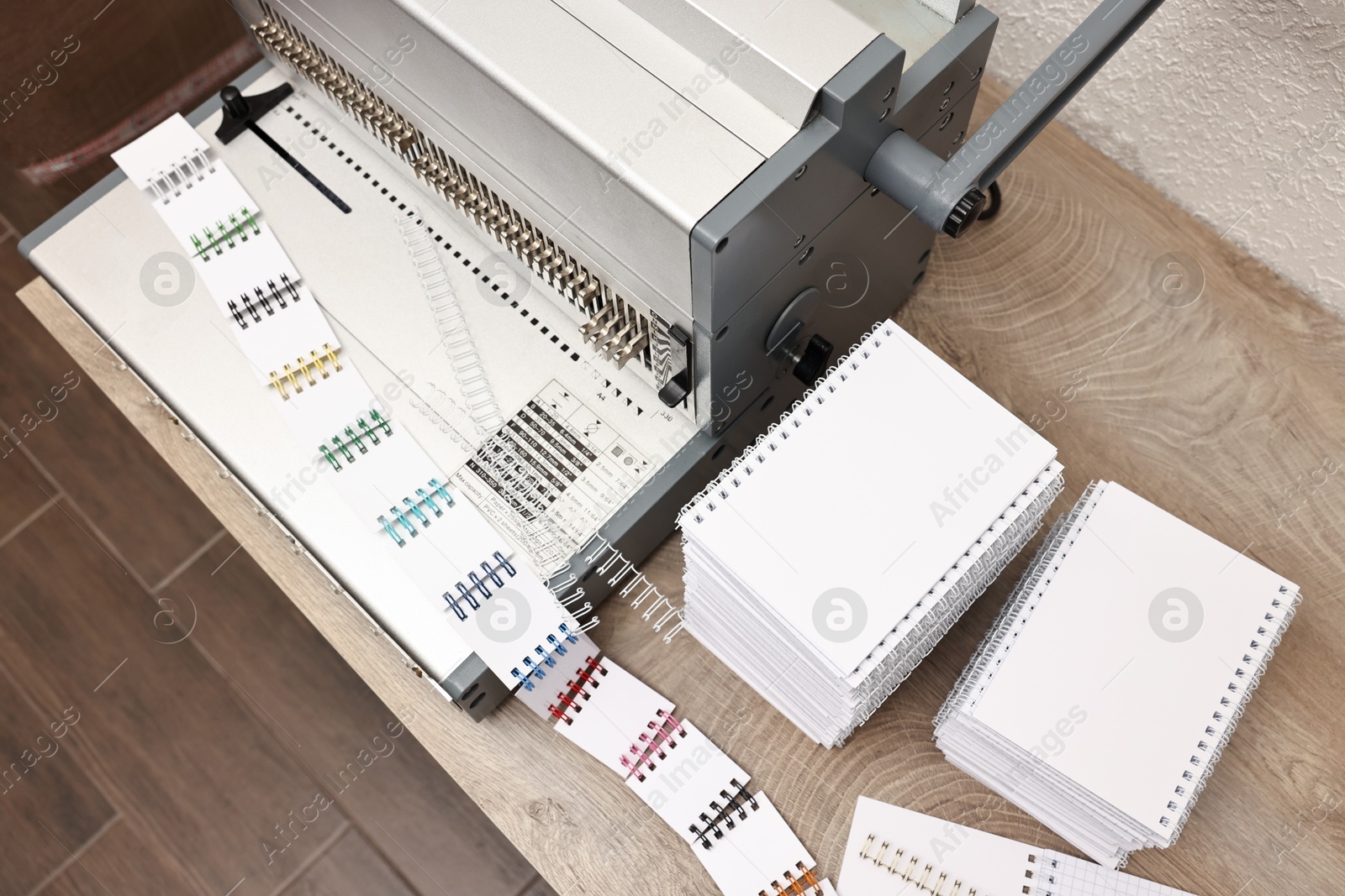 Photo of Modern binding machine, stacks of notebooks and double loop wire binding spines on wooden table indoors, above view