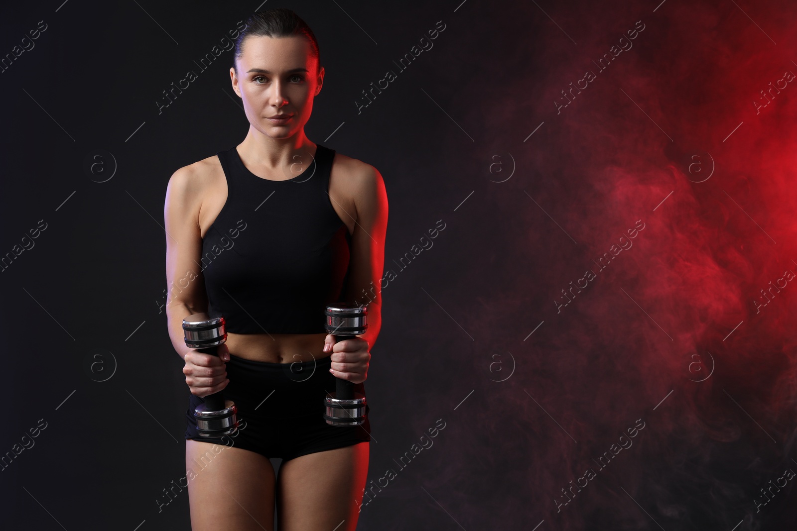 Photo of Woman exercising with dumbbells in smoke on dark background, space for text