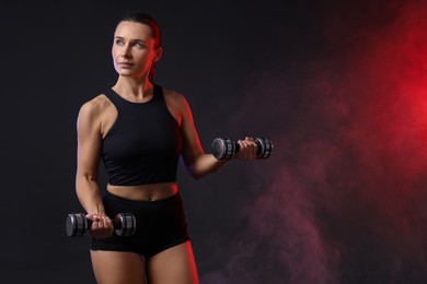 Woman exercising with dumbbells in smoke on dark background, space for text