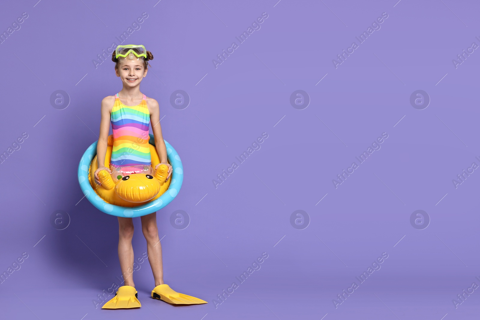 Photo of Happy girl in beachwear with diving mask, flippers and inflatable ring on purple background, space for text