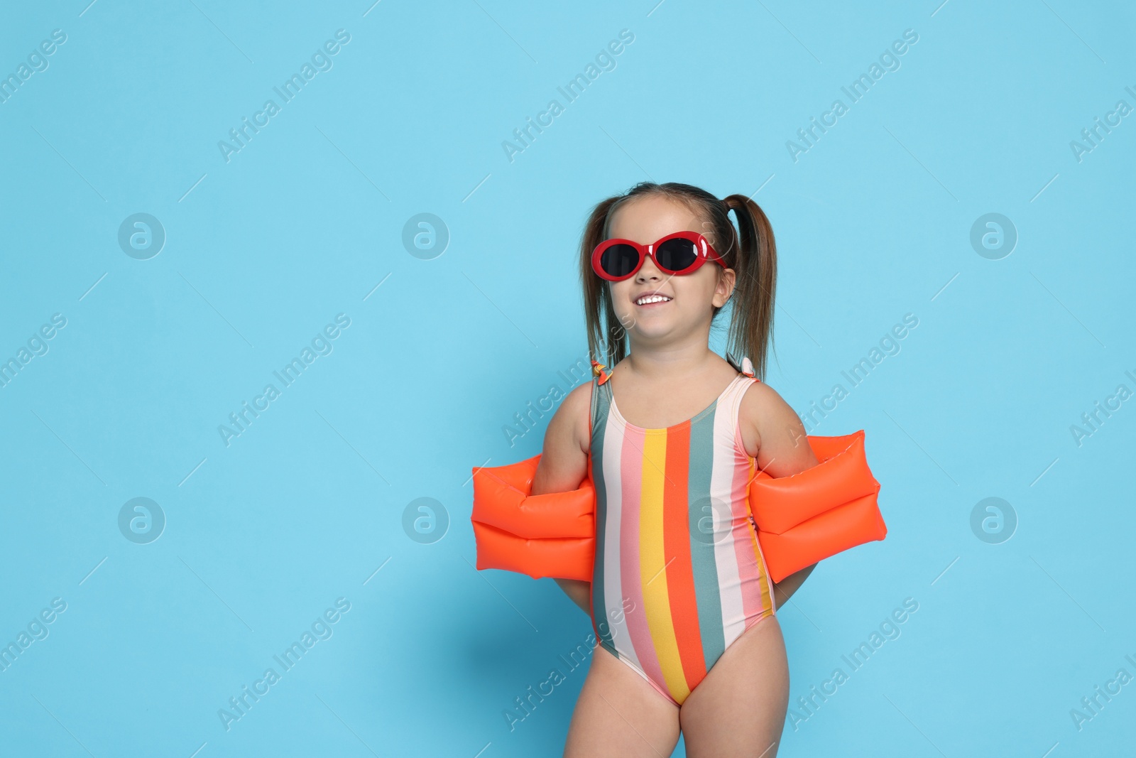 Photo of Cute little girl in swimsuit with swim armbands on light blue background. Space for text