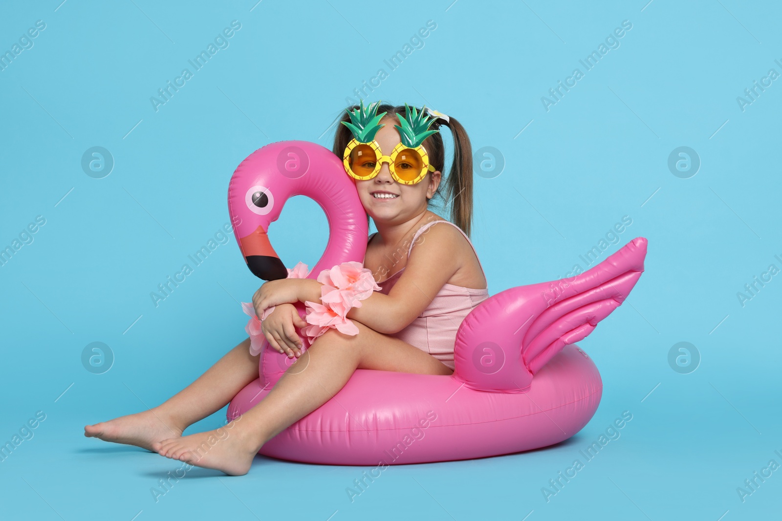 Photo of Cute little girl in swimsuit with funny sunglasses and inflatable ring on light blue background