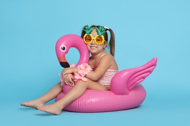 Cute little girl in swimsuit with funny sunglasses and inflatable ring on light blue background