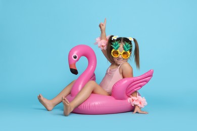 Cute little girl in swimsuit with funny sunglasses and inflatable ring on light blue background