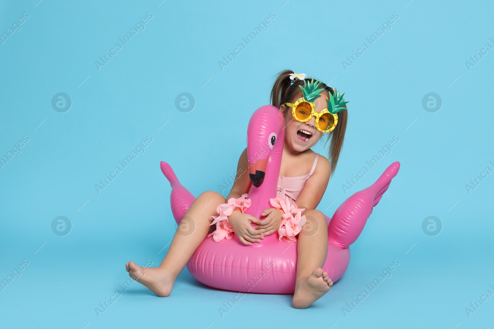 Photo of Cute little girl in swimsuit with funny sunglasses and inflatable ring on light blue background