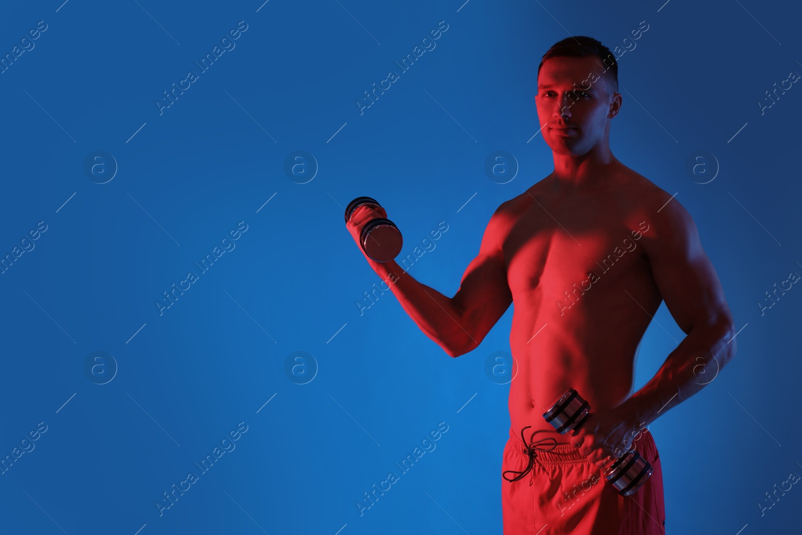 Photo of Man exercising with dumbbells in red light on blue background, space for text