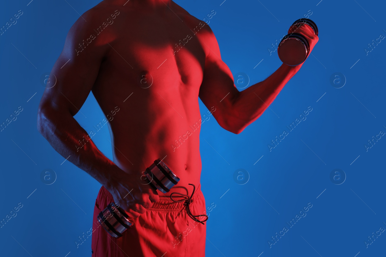 Photo of Man exercising with dumbbells in red light on blue background, closeup