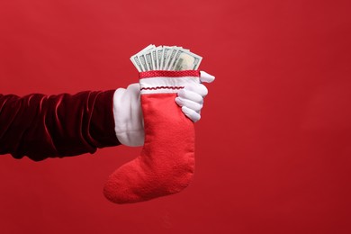 Photo of Santa Claus holding stocking with dollar banknotes on red background, closeup. Space for text