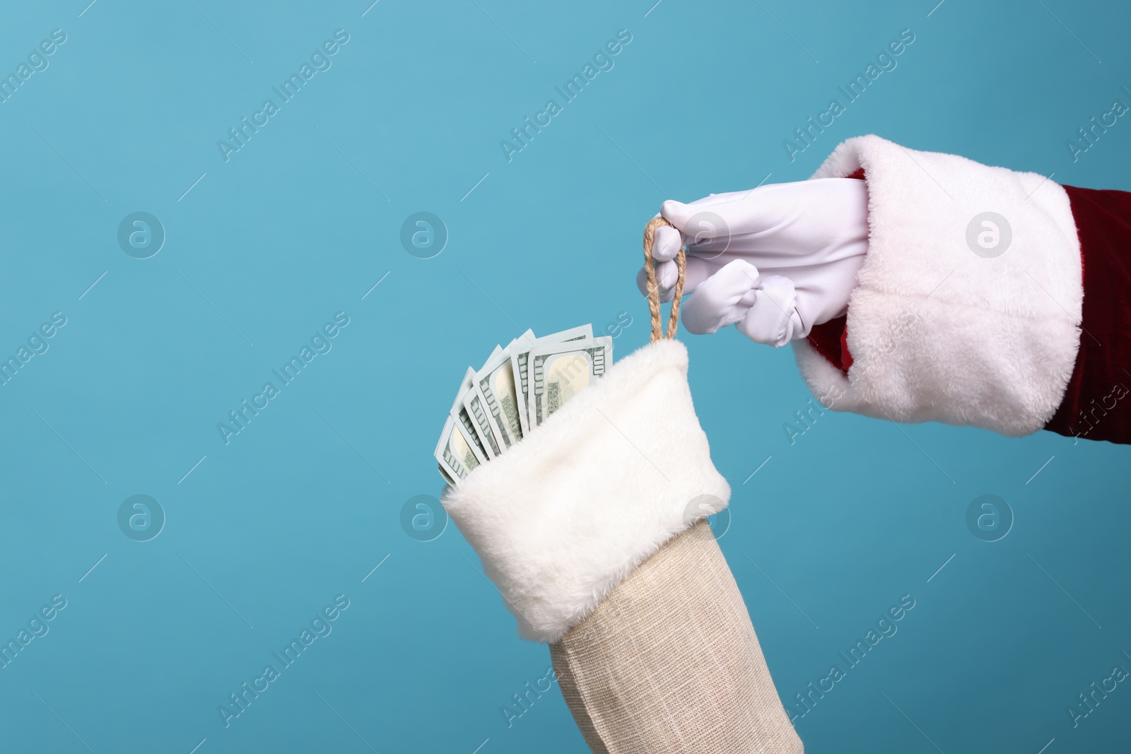 Photo of Santa Claus holding stocking with dollar banknotes on light blue background, closeup. Space for text